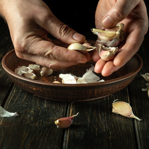 The cook cleans the garlic with his hands in a vintage plate on the kitchen table before preparing a vegetarian meal. Tasty recipe concept on black background.